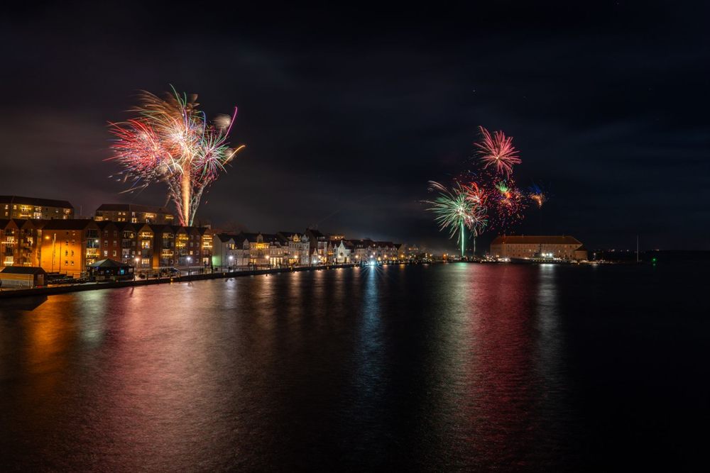 Fireworks over a harbor town with reflections on the water at night